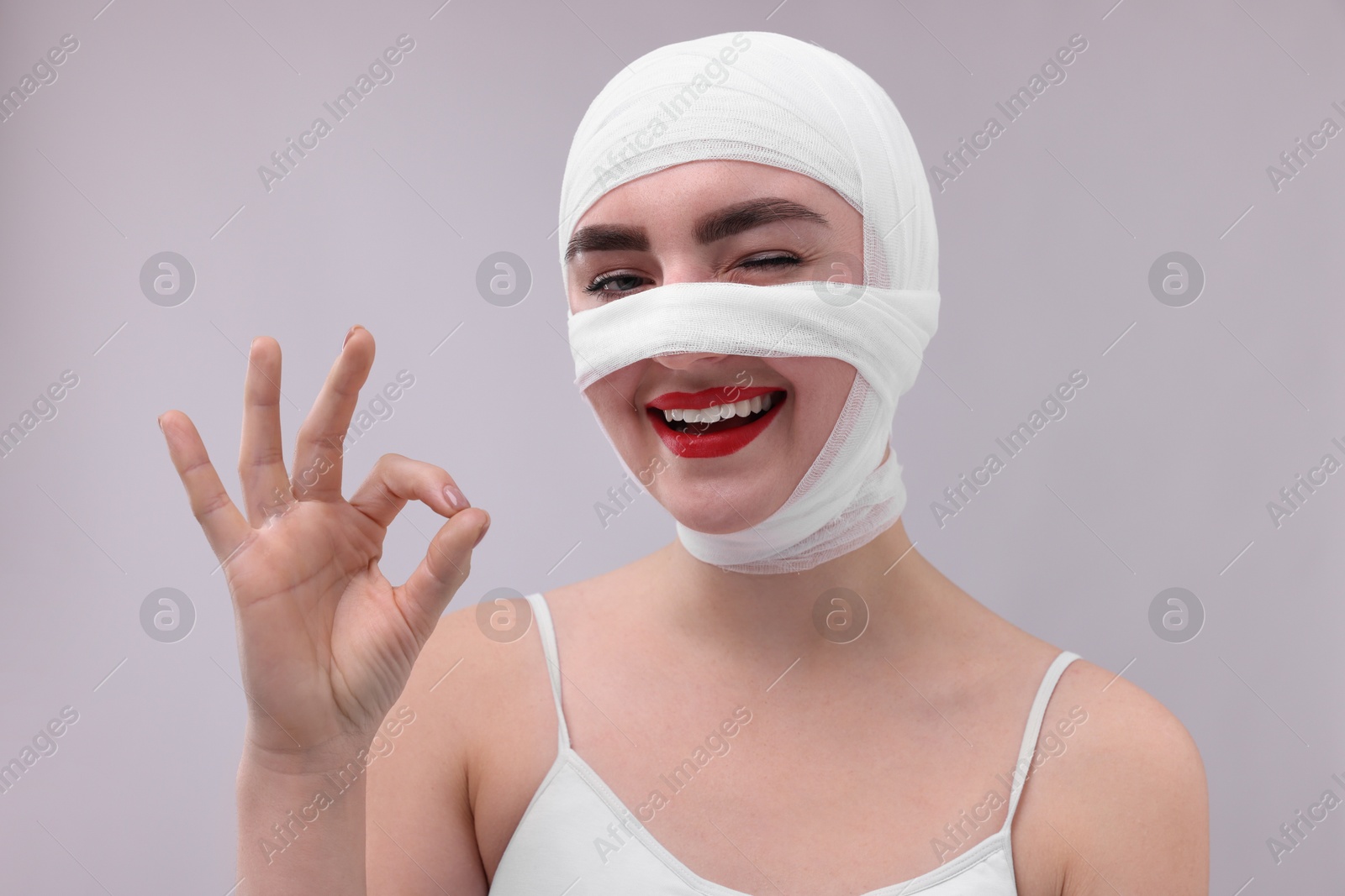 Photo of Woman with nose wrapped in medical bandage after plastic surgery operation showing ok gesture on grey background