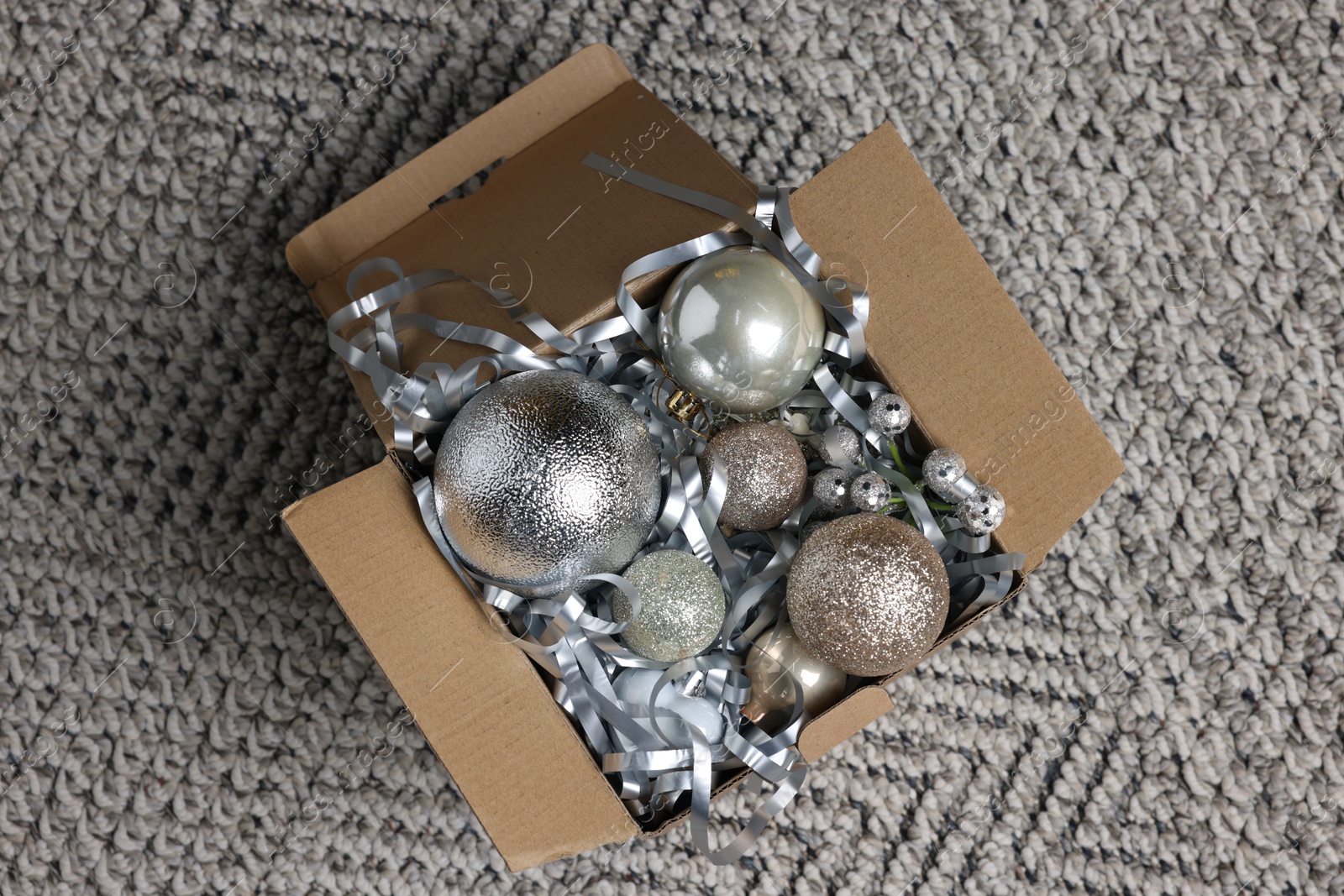 Photo of Shiny tinsel and Christmas balls in cardboard box on carpet, top view