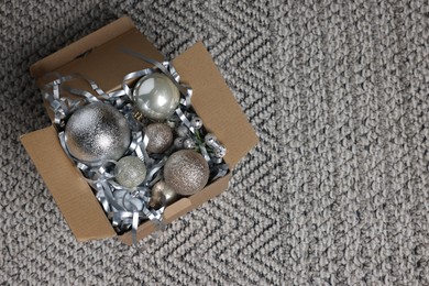 Photo of Shiny tinsel and Christmas balls in cardboard box on carpet, top view. Space for text