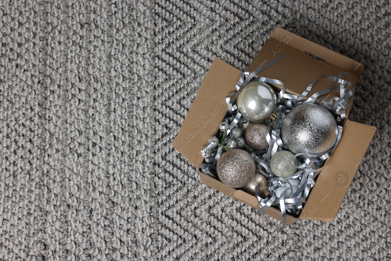 Photo of Shiny tinsel and Christmas balls in cardboard box on carpet, top view. Space for text