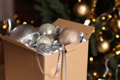 Photo of Shiny tinsel and Christmas balls in cardboard box against blurred lights, closeup