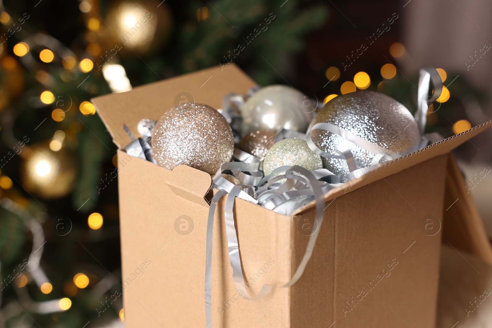 Photo of Shiny tinsel and Christmas balls in cardboard box against blurred lights, closeup