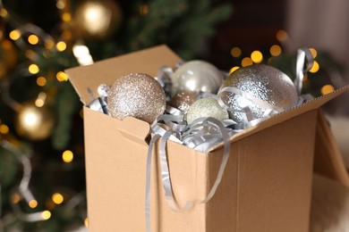 Photo of Shiny tinsel and Christmas balls in cardboard box against blurred lights, closeup