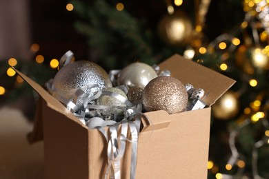 Photo of Shiny tinsel and Christmas balls in cardboard box against blurred lights, closeup