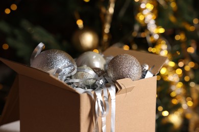 Photo of Shiny tinsel and Christmas balls in cardboard box against blurred lights, closeup