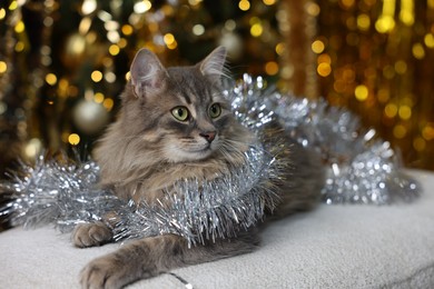 Photo of Cute cat with shiny tinsel on pouf against blurred lights. Christmas atmosphere