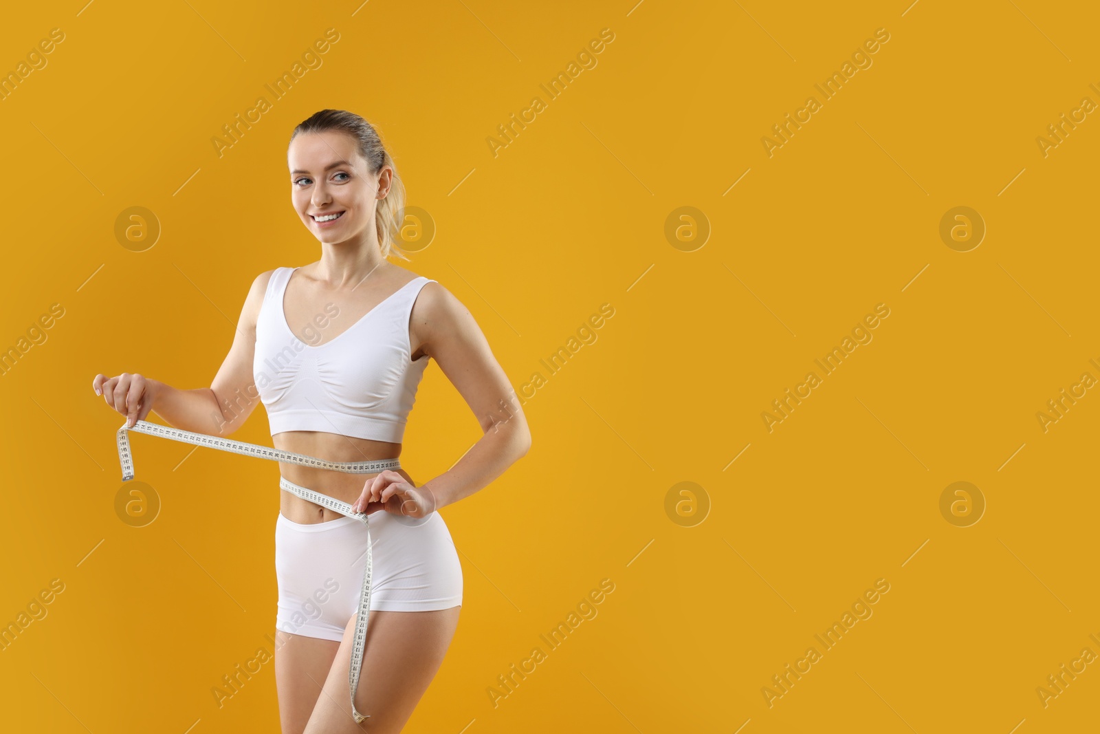 Photo of Weight loss. Happy woman measuring her slim waist with tape on yellow background, space for text