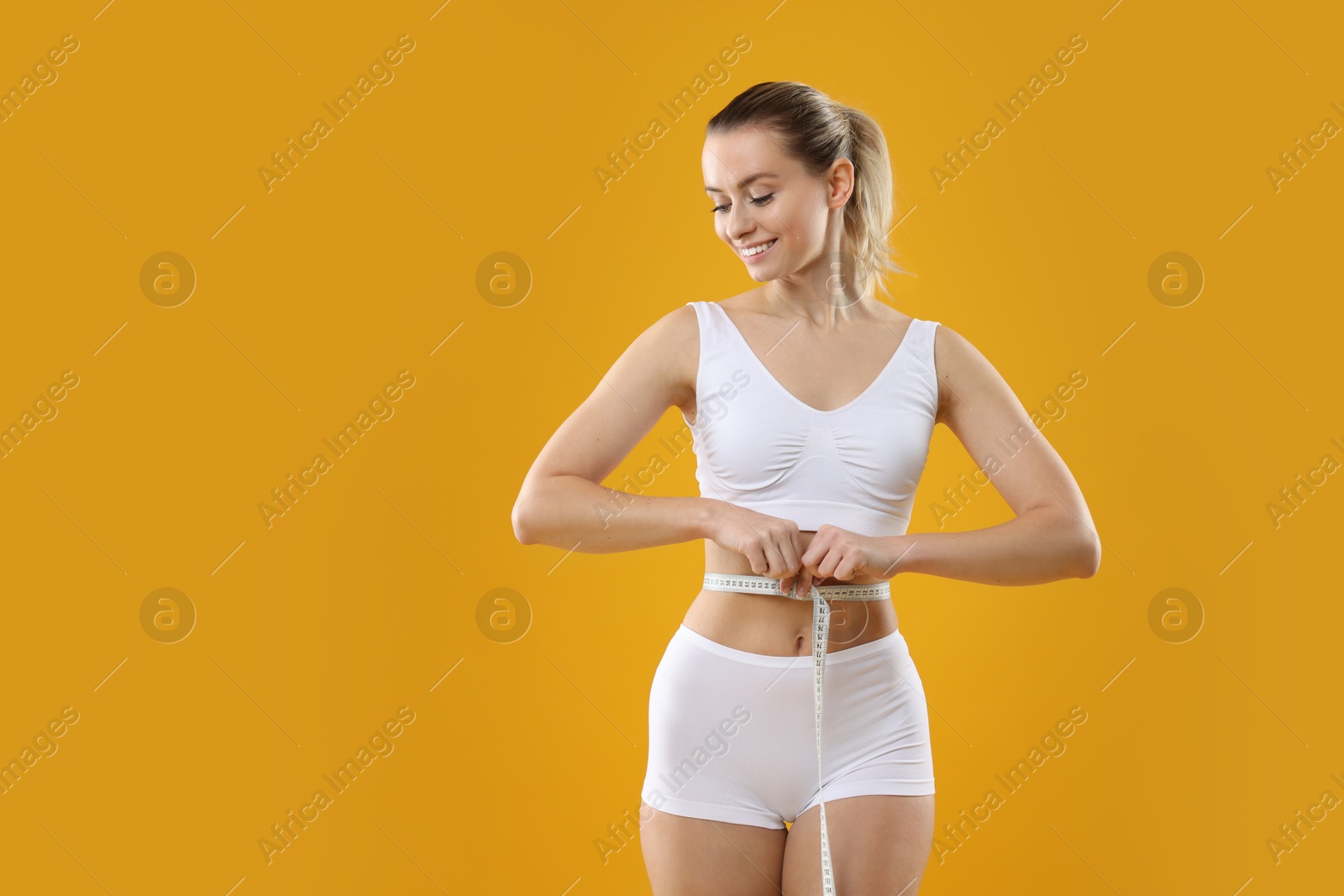 Photo of Weight loss. Happy woman measuring her slim waist with tape on yellow background, space for text