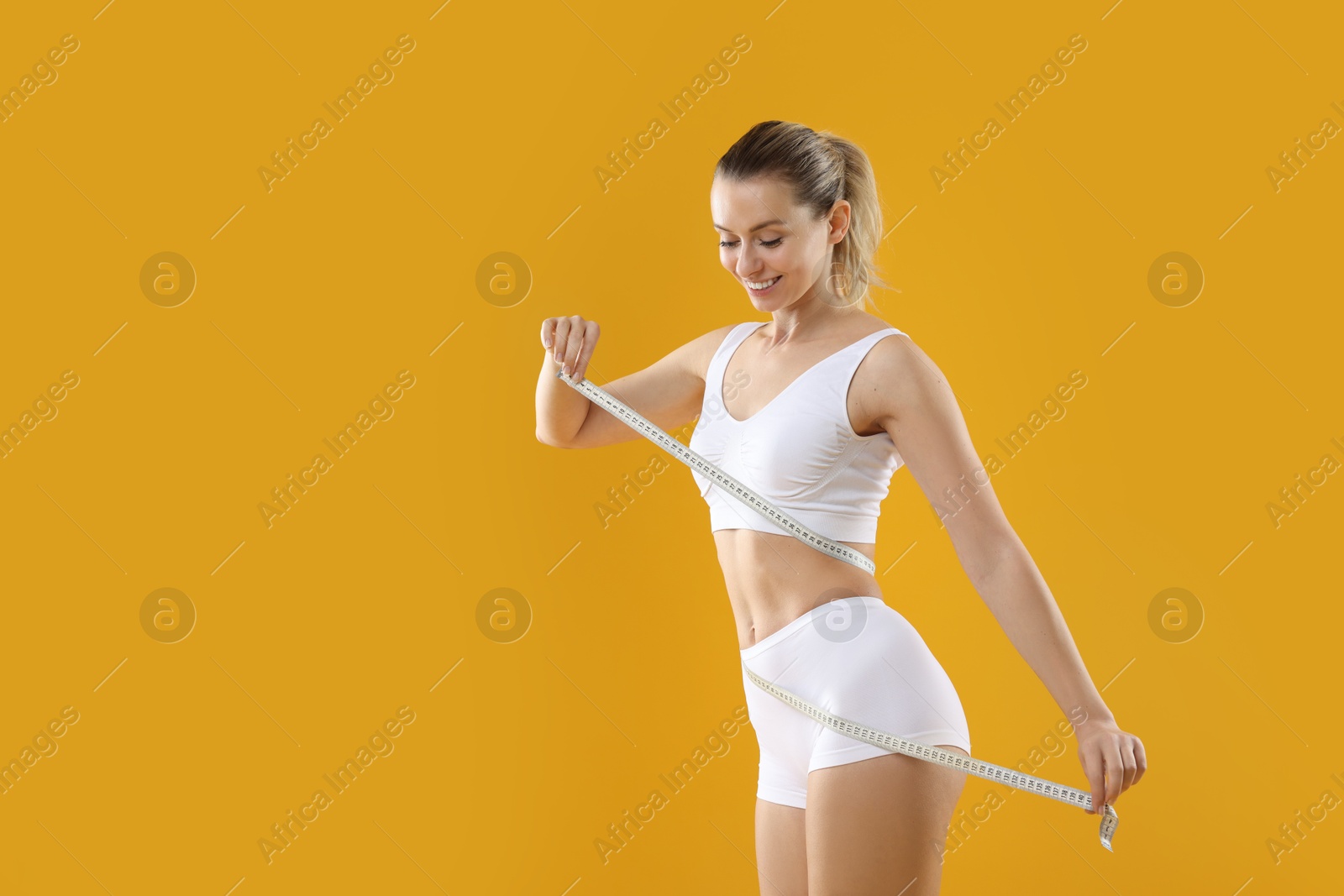 Photo of Weight loss. Happy woman measuring her slim waist with tape on yellow background, space for text