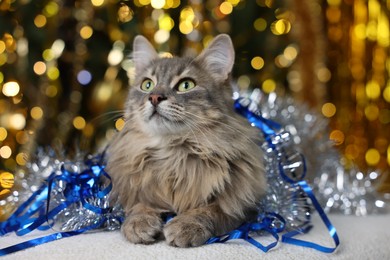 Photo of Cute cat with shiny tinsels on pouf against blurred lights. Christmas atmosphere