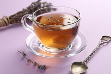 Photo of Aromatic lavender tea in glass cup, spoon and dry flowers on pink background, closeup