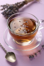 Photo of Aromatic lavender tea in glass cup, spoon and dry flowers on pink background, closeup