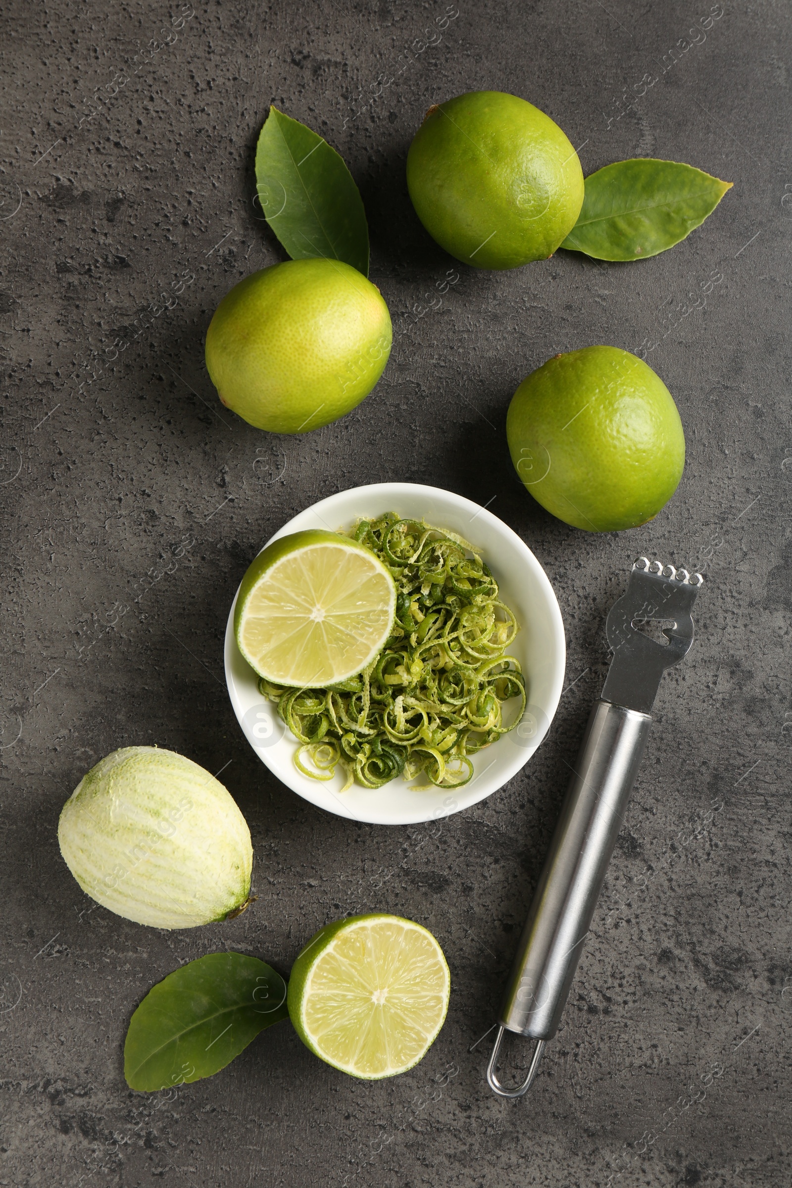 Photo of Lime zest, fresh fruits and zester on grey textured table