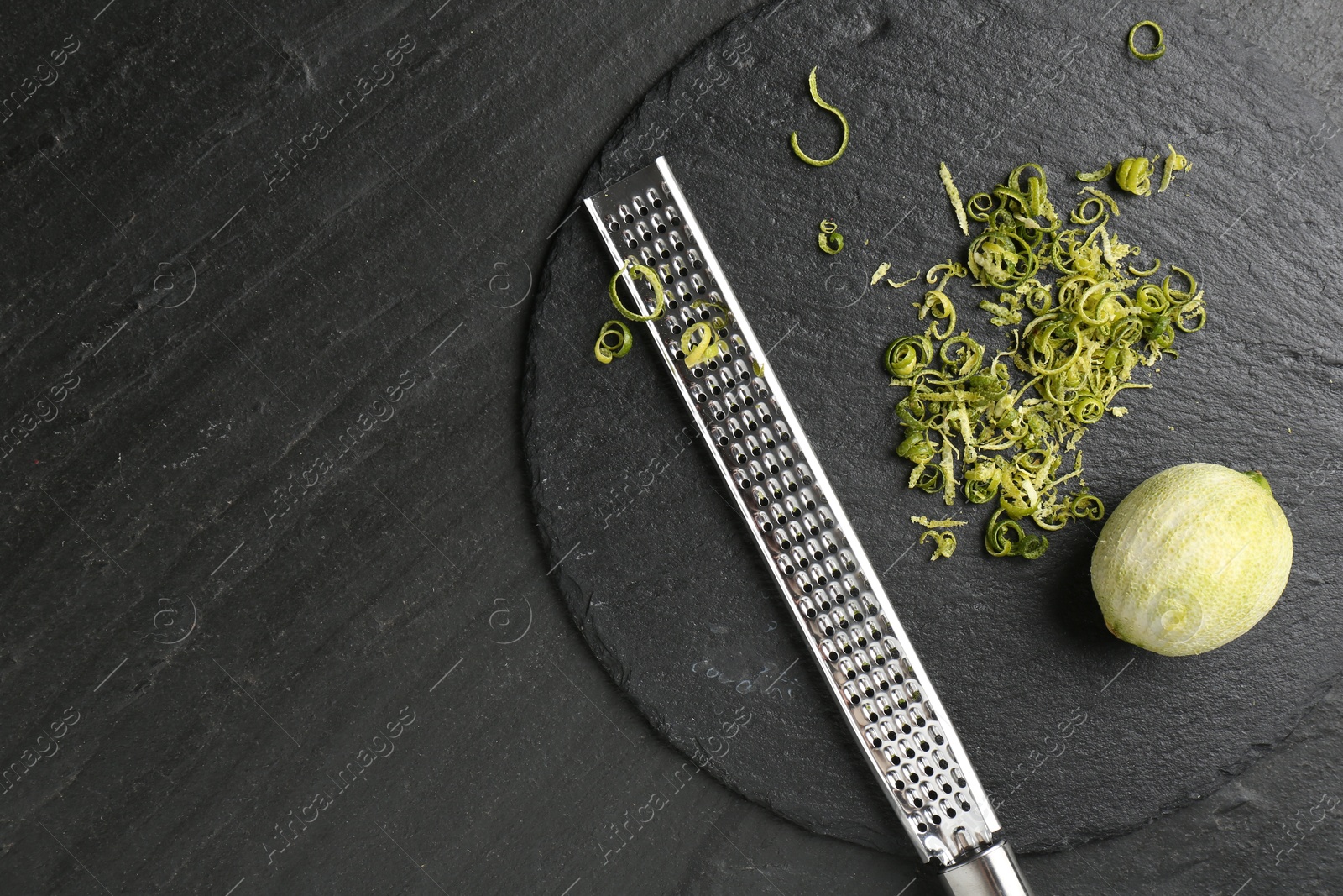 Photo of Lime zest, grater and peeled fruit on dark textured table, top view. Space for text