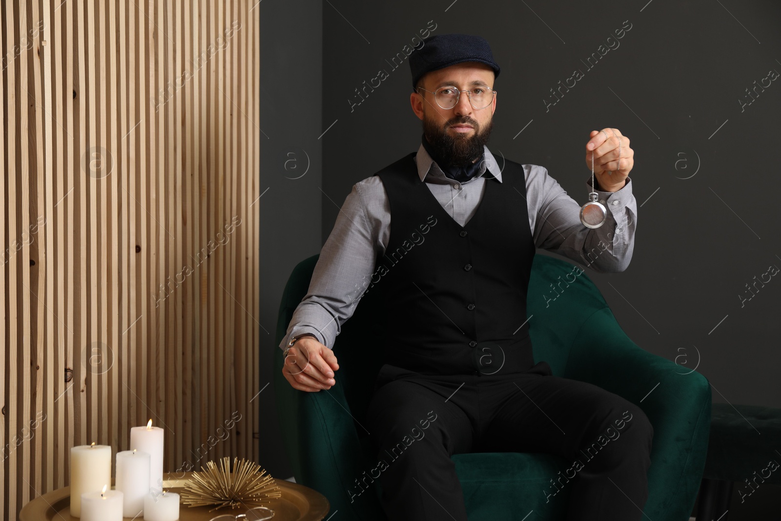 Photo of Hypnosis session. Man with vintage pocket watch in armchair near grey wall indoors