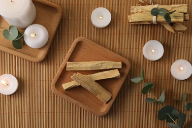 Photo of Palo santo sticks, eucalyptus branches and burning candles on bamboo mat, flat lay