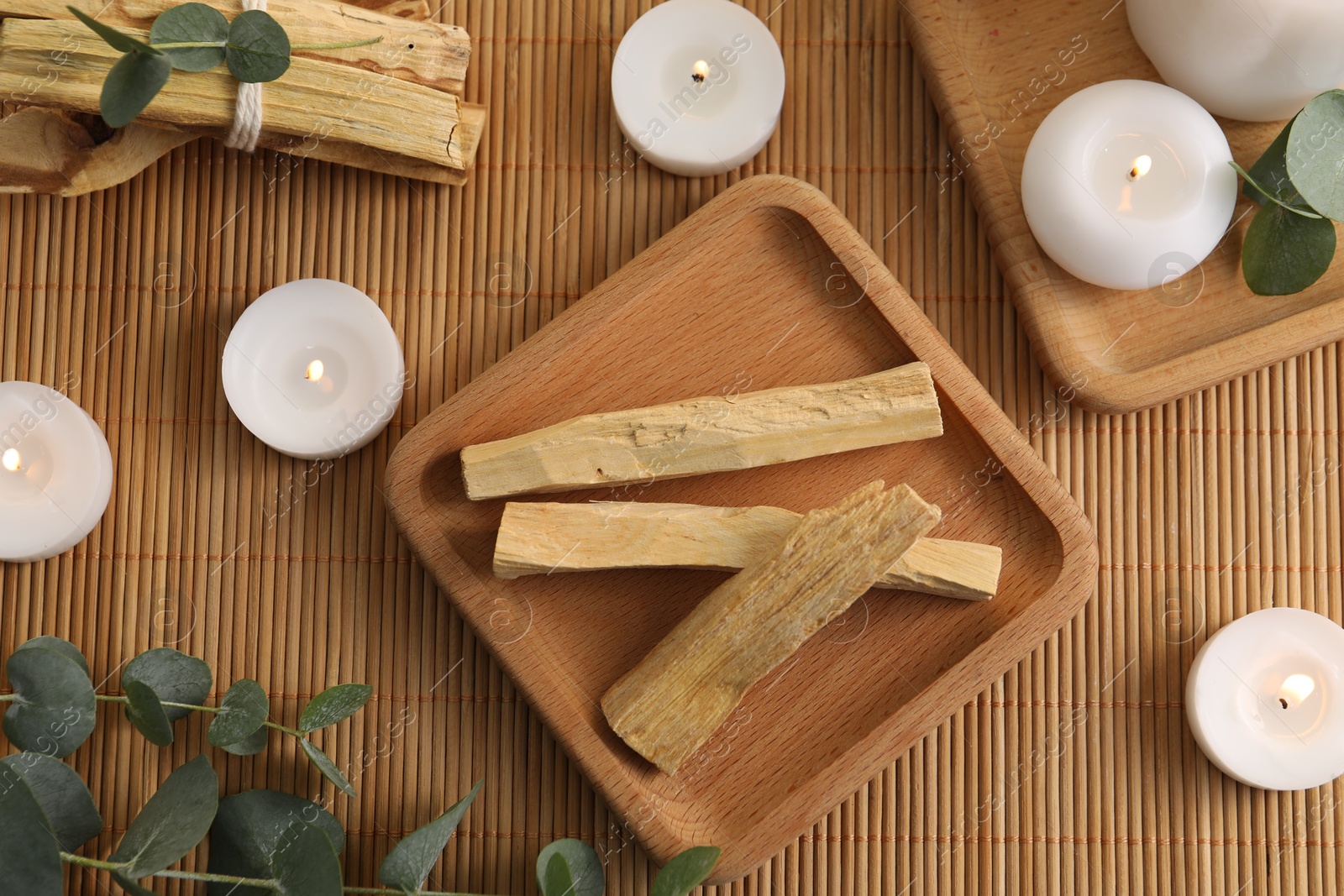 Photo of Palo santo sticks, eucalyptus branches and burning candles on bamboo mat, flat lay