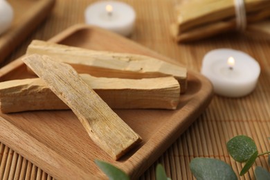 Photo of Palo santo sticks, eucalyptus branches and burning candles on bamboo mat, closeup