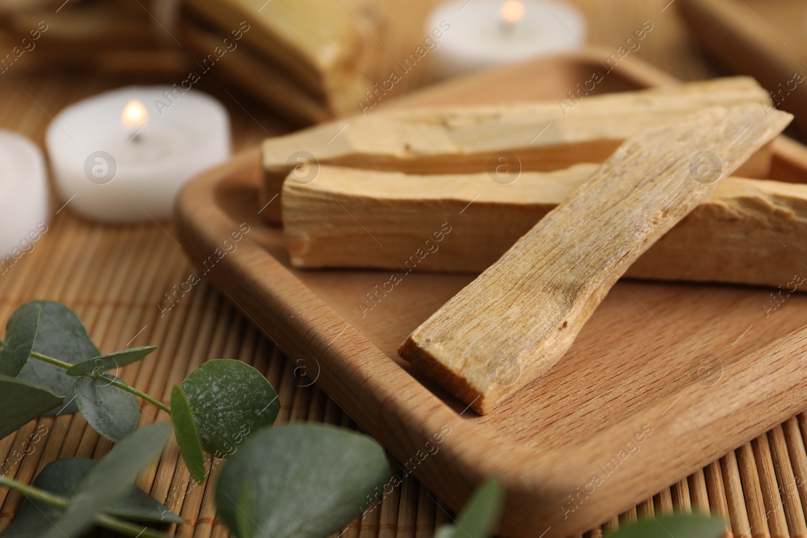 Photo of Palo santo sticks, eucalyptus branches and burning candles on bamboo mat, closeup