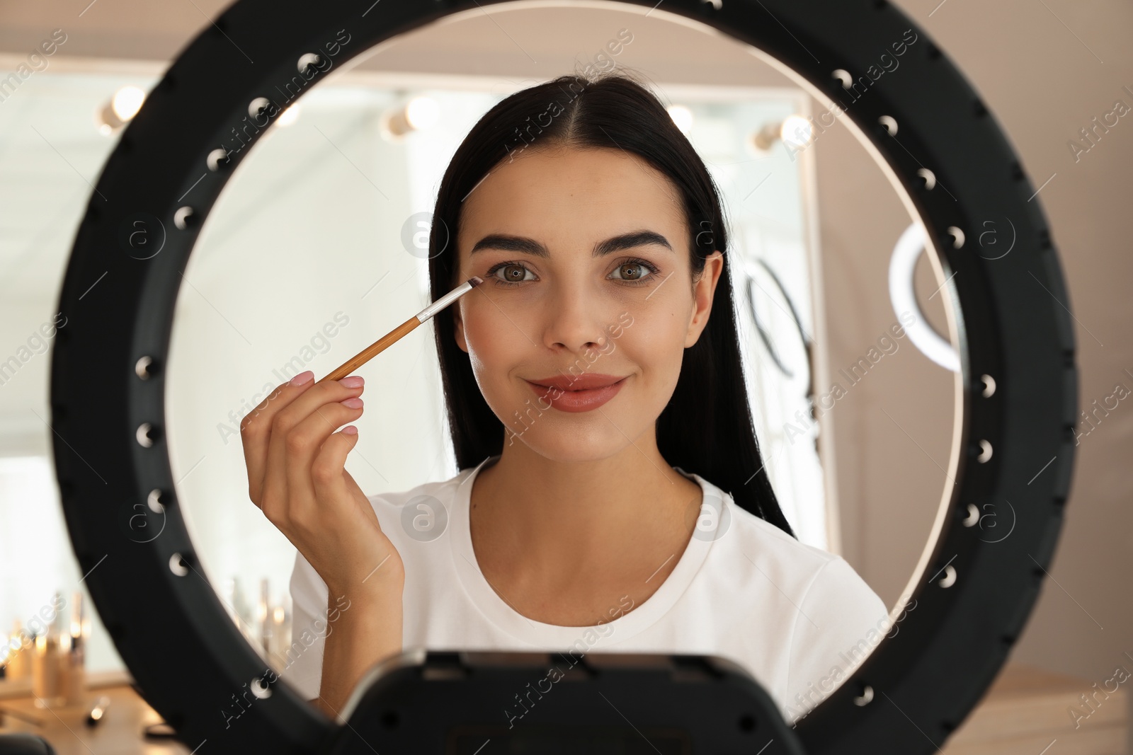 Photo of Beautiful young woman applying eye shadow with brush indoors, view through ring lamp