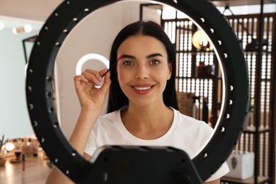 Photo of Beautiful young woman brushing eyebrows indoors, view through ring lamp