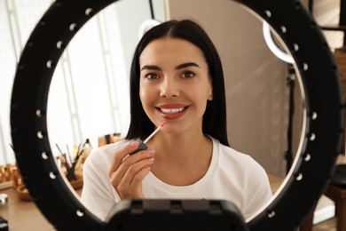 Photo of Beautiful young woman applying liquid lipstick indoors, view through ring lamp