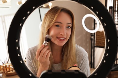 Photo of Beautiful young woman applying face powder with brush indoors, view through ring lamp