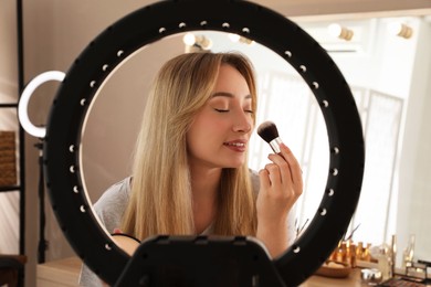 Photo of Beautiful young woman applying face powder with brush indoors, view through ring lamp