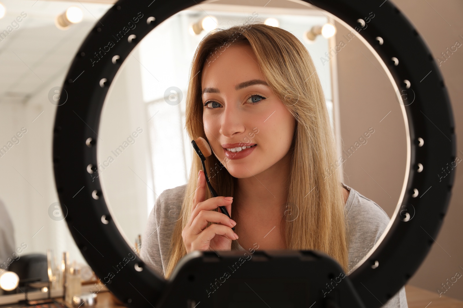 Photo of Beautiful young woman applying foundation with brush indoors, view through ring lamp