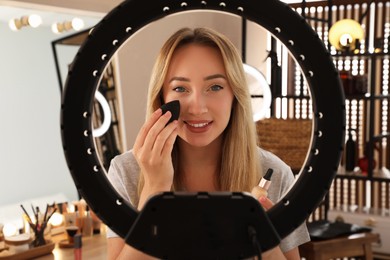 Photo of Beautiful young woman applying foundation with beauty blender indoors, view through ring lamp