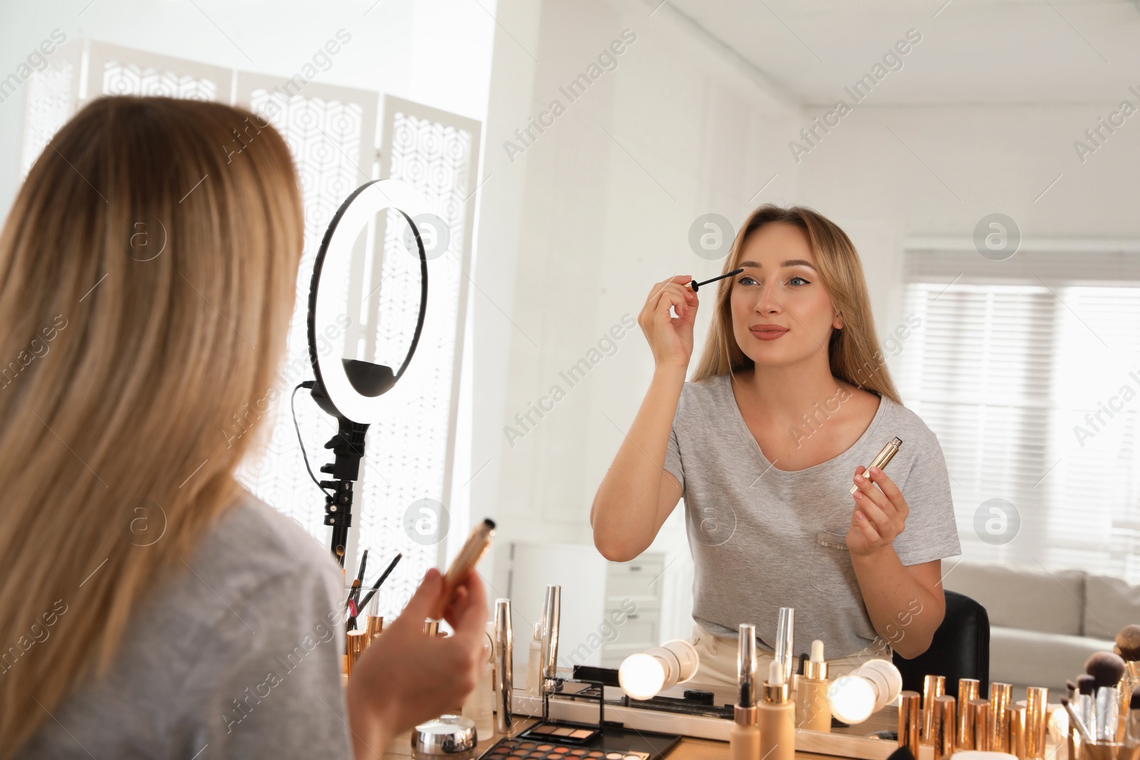 Photo of Beautiful young woman applying makeup at table with mirror and ring lamp