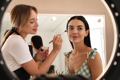 Professional makeup artist working with beautiful young woman in salon, view through ring lamp