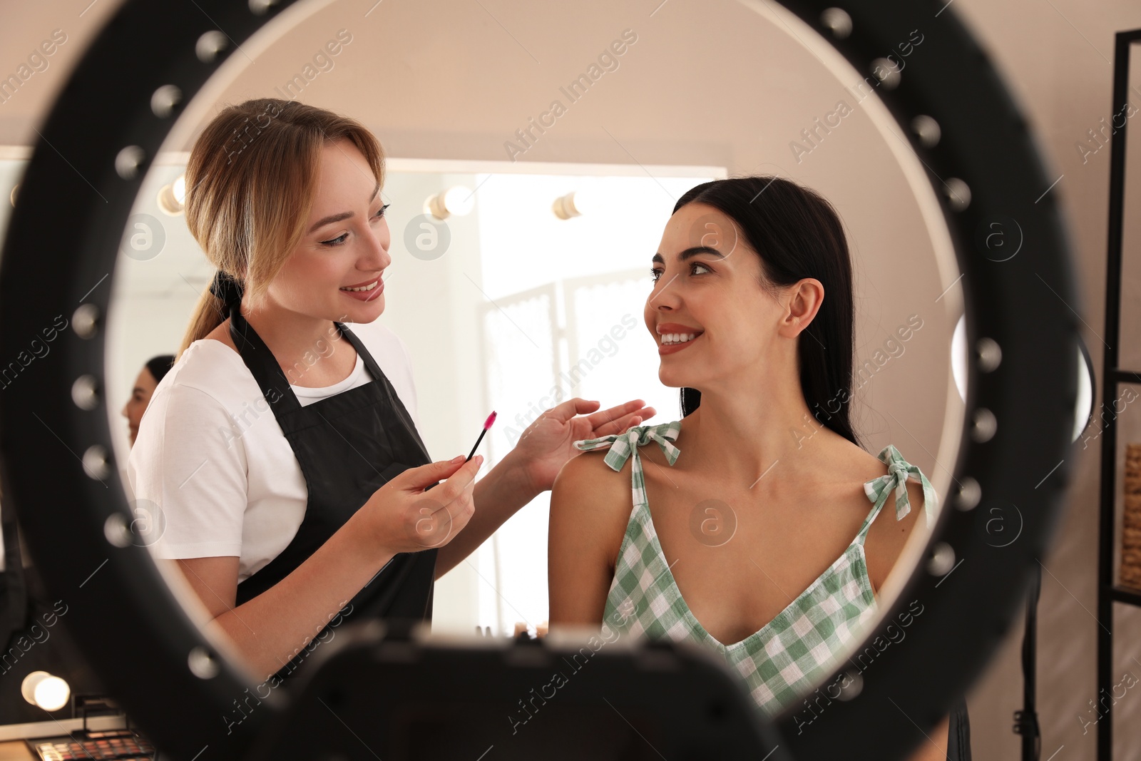 Photo of Professional makeup artist working with beautiful young woman in salon, view through ring lamp