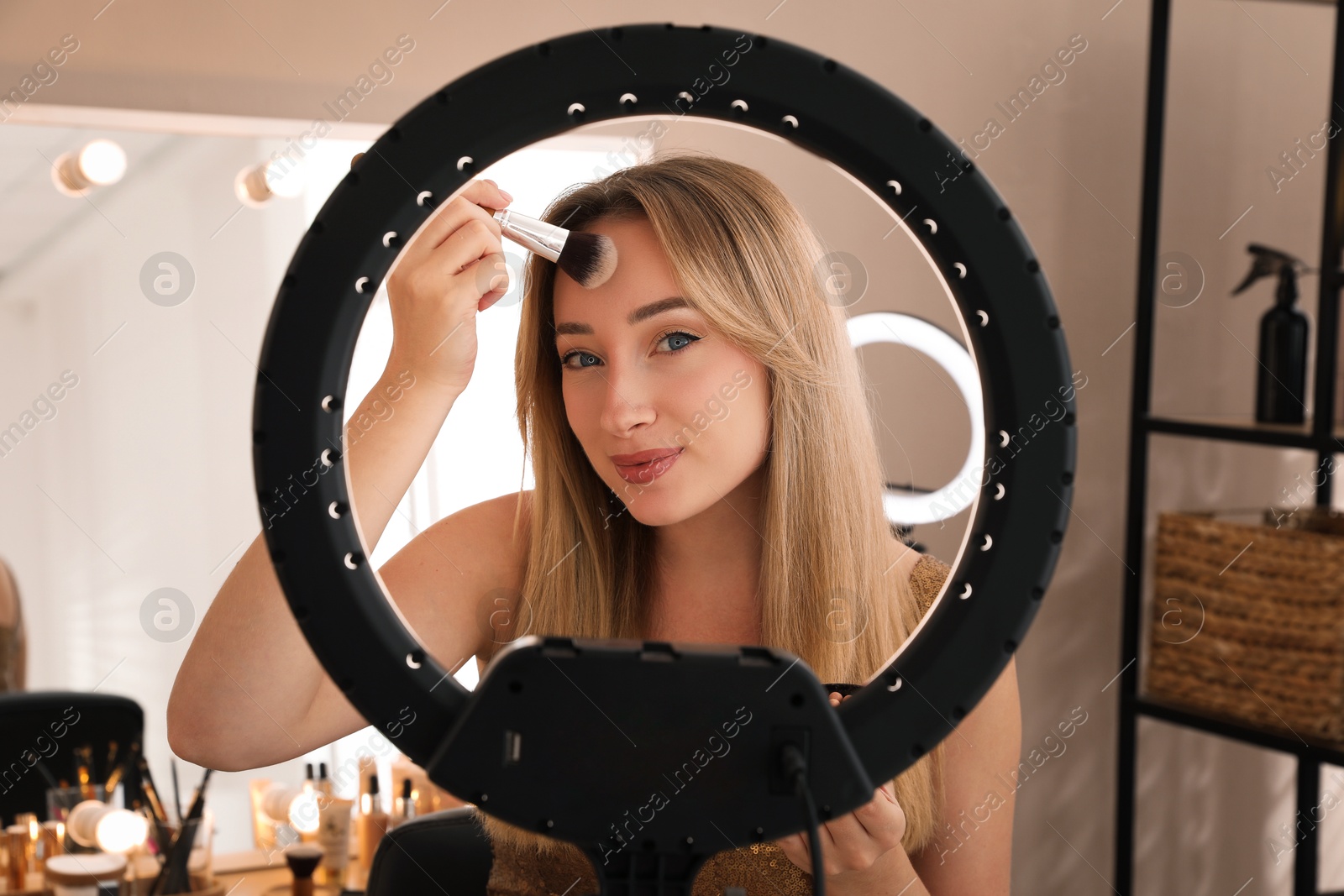 Photo of Beautiful young woman applying makeup indoors, view through ring lamp