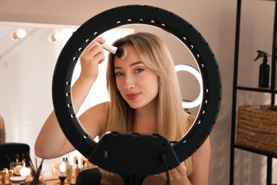 Photo of Beautiful young woman applying makeup indoors, view through ring lamp