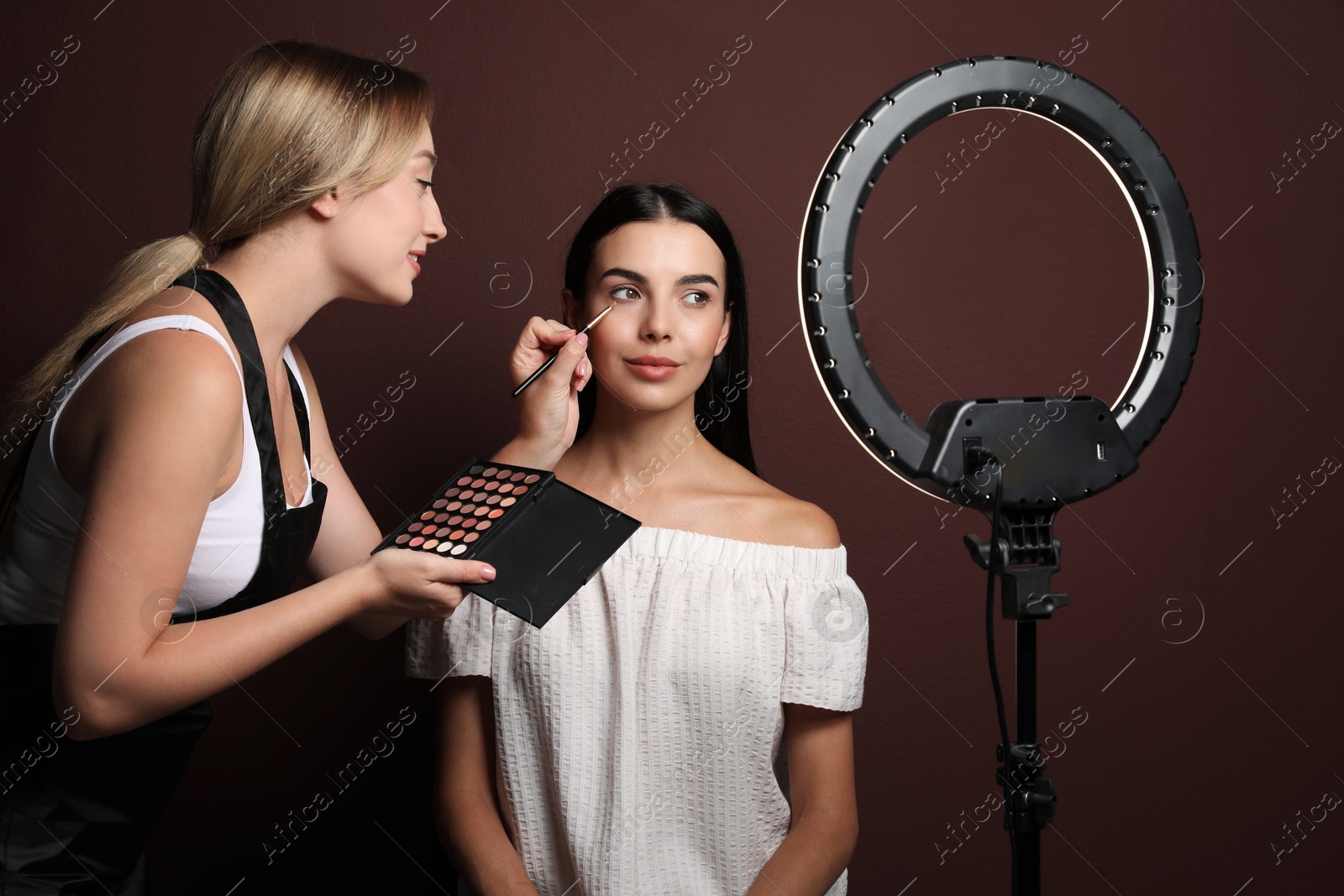 Photo of Professional makeup artist working with beautiful young woman against brown background. Using ring lamp