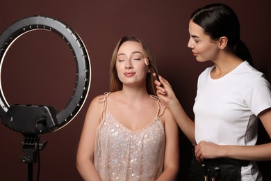 Professional makeup artist working with beautiful young woman against brown background. Using ring lamp