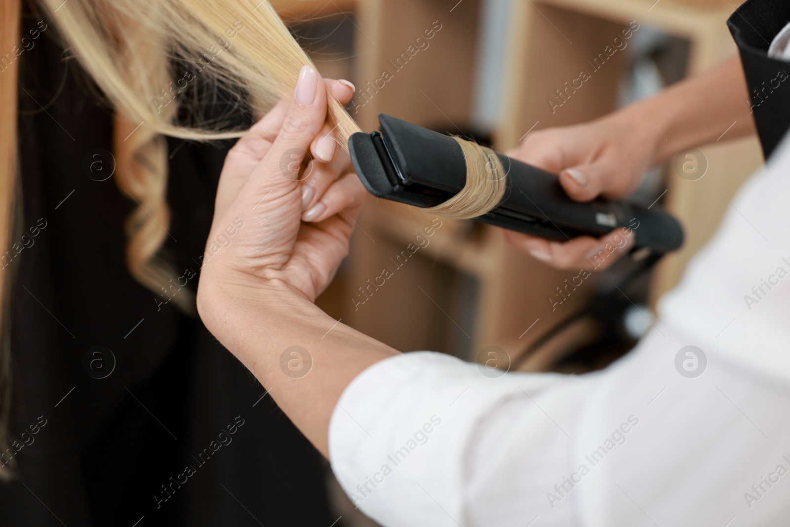 Photo of Hairdresser curling woman's hair with flat iron in salon, closeup