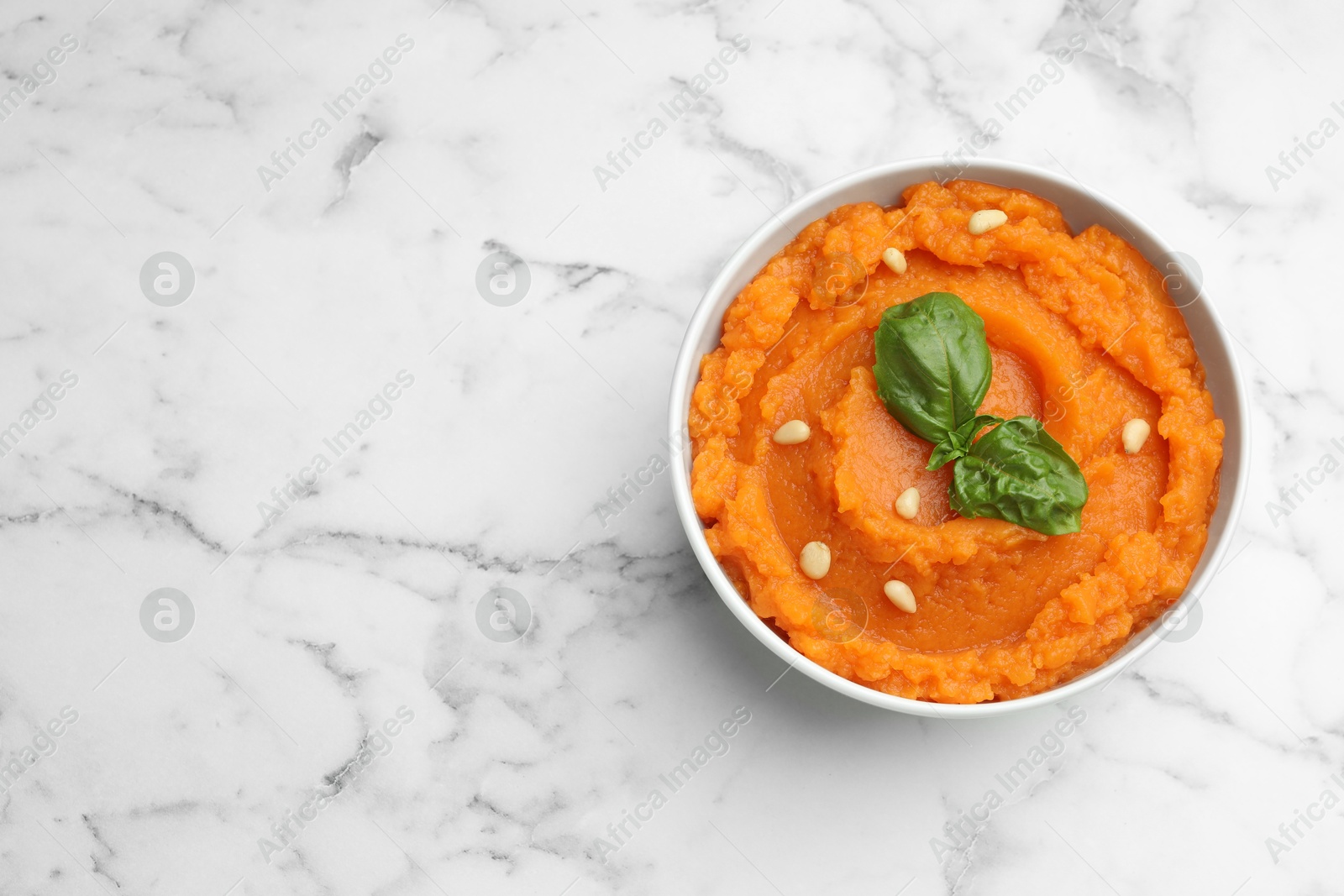 Photo of Tasty mashed sweet potato with basil and seeds in bowl on white marble table, top view. Space for text