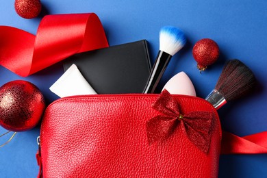 Photo of Christmas gift. Decorative cosmetics in red makeup bag and festive decor on blue background, flat lay