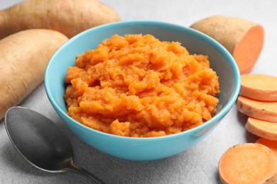 Photo of Tasty mashed sweet potato in bowl, spoon and fresh vegetables on gray table, closeup