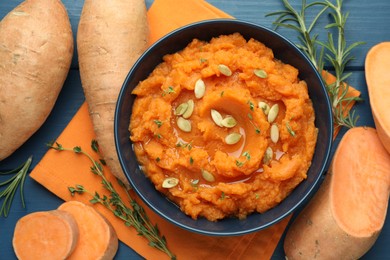 Photo of Tasty mashed sweet potato with pumpkin seeds in bowl, fresh vegetables, rosemary and thyme on blue wooden table, flat lay