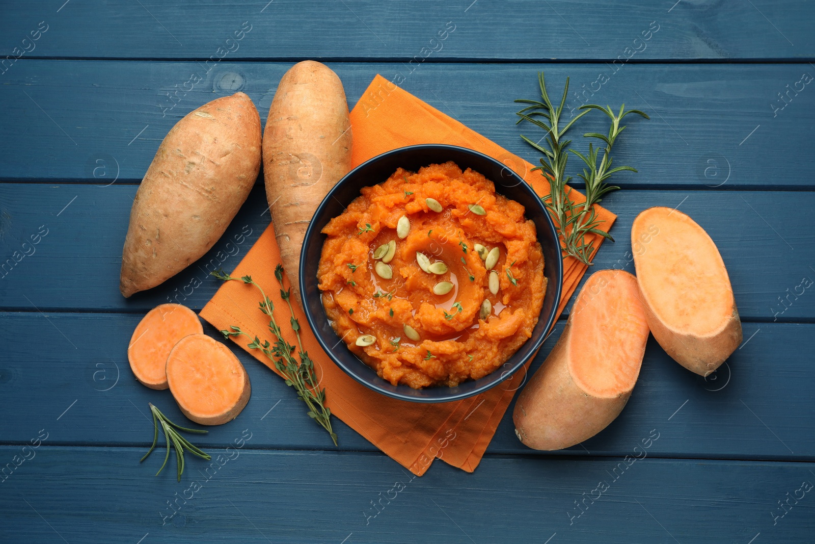 Photo of Tasty mashed sweet potato with pumpkin seeds in bowl, fresh vegetables, rosemary and thyme on blue wooden table, flat lay