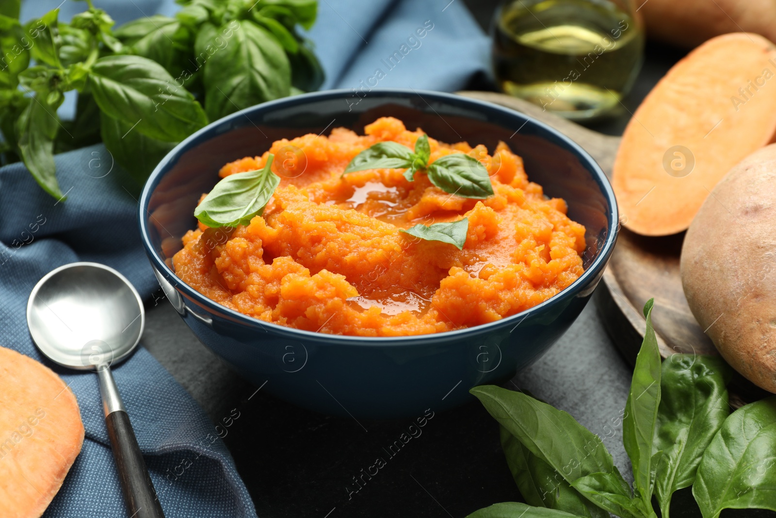 Tasty mashed sweet potato with basil in bowl, fresh vegetables and spoon on table, closeup Photo of Tasty mashed sweet potato with basil in bowl, fresh vegetables and spoon on table, closeup