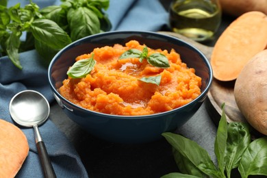 Photo of Tasty mashed sweet potato with basil in bowl, fresh vegetables and spoon on table, closeup