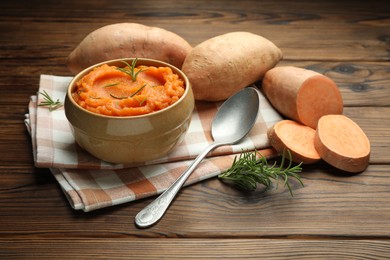 Photo of Tasty mashed sweet potato with rosemary in bowl, fresh vegetables and spoon on wooden table, closeup