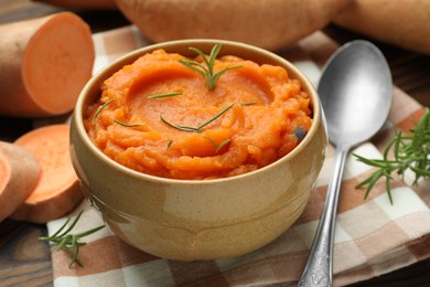 Photo of Tasty mashed sweet potato with rosemary in bowl, fresh vegetables and spoon on table, closeup