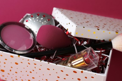 Christmas gift box with decorative cosmetics on dark pink background, closeup