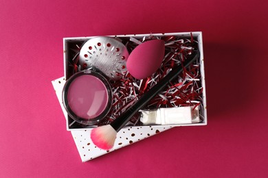 Photo of Christmas gift box with decorative cosmetics on dark pink background, top view