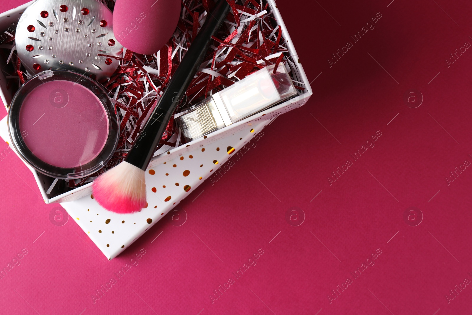 Photo of Christmas gift box with decorative cosmetics on dark pink background, top view. Space for text